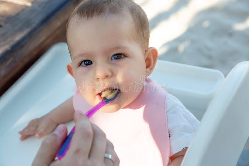 Un peque?o viejo beb? de ocho meses con placer come las gachas de avena de una cuchara Se sienta en la playa en una silla de los fotografía de archivo Un peque?o viejo beb? de ocho meses con placer come las gachas de avena de una cuchara Se sienta en la playa en una silla de los fotografía de archivo