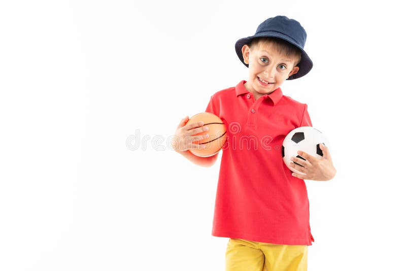 Un petit garçon au panama, maillot jaune, short rouge et baskets blanches tient des stands de basket et de football photographie stock Un petit garçon au panama, maillot jaune, short rouge et baskets blanches tient des stands de basket et de football photographie stock