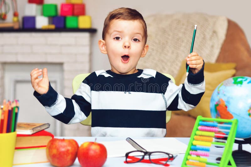 Un petit garçon d'âge préscolaire surpris s'assoit à une table avec un livre un globe un cahier et des crayons image libre de droits