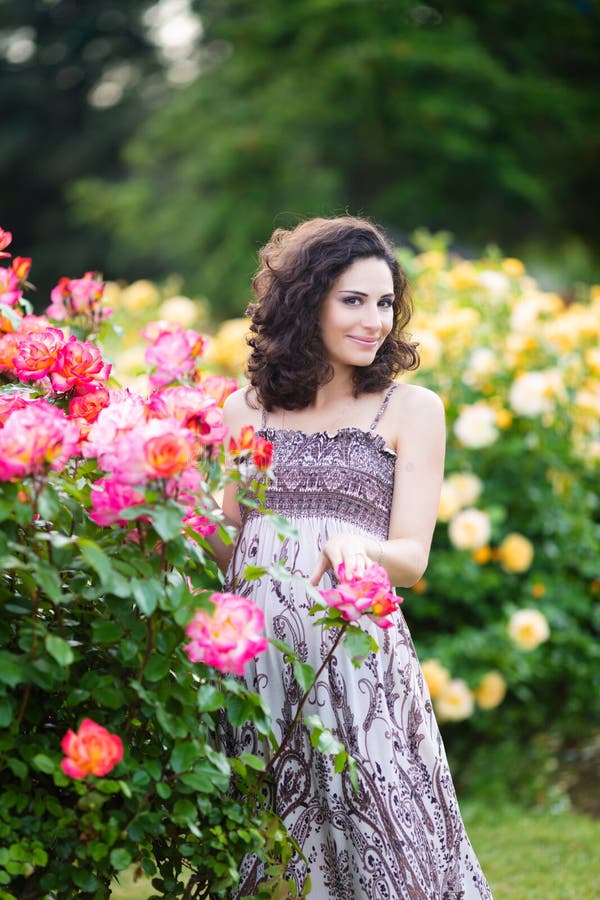 Un portrait vertical de jeune femme caucasienne avec les cheveux bouclés bruns foncés près des rosiers roses, regardant vers sa g photographie stock Un portrait vertical de jeune femme caucasienne avec les cheveux bouclés bruns foncés près des rosiers roses, regardant vers sa g photographie stock