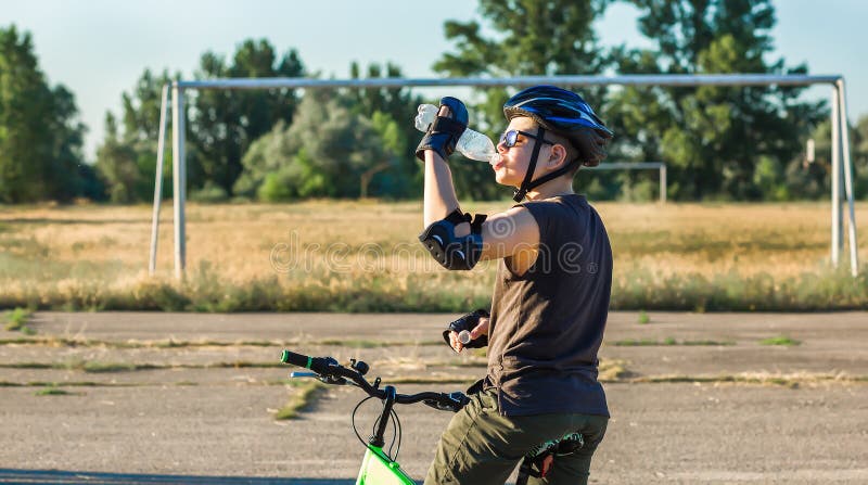 A boy in a safety helmet on a bicycle drinks water from a plastic bottle. Thirst. Cycling. Sports stadium. Active lifestyle concept. A boy in a safety helmet on a bicycle drinks water from a plastic bottle. Thirst. Cycling. Sports stadium. Active lifestyle concept