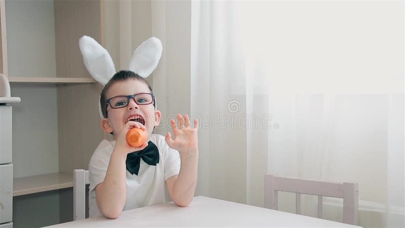 Un ragazzo con le orecchie del coniglietto sulla sua testa che si siede ad una tavola bianca che mangia una carota