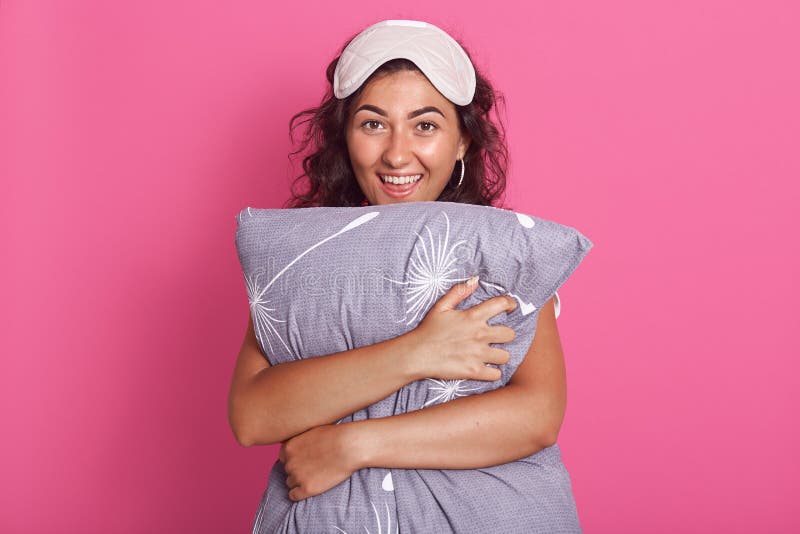 Un retrato de feliz joven en pijama y mascarilla dormido abrazando almohada sobre fondo rosa, parece sonreír directamente foto de archivo libre de regalías Un retrato de feliz joven en pijama y mascarilla dormido abrazando almohada sobre fondo rosa, parece sonreír directamente foto de archivo libre de regalías