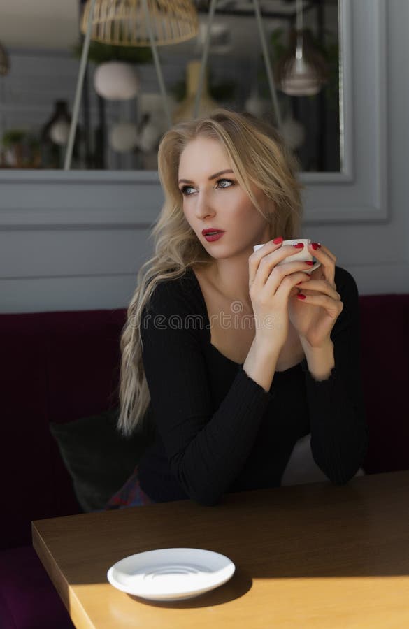 Un rubio de pelo largo hermoso se sienta y descansa en una tabla en un café, sosteniendo una taza de café y mirando hacia fuera l imagenes de archivo Un rubio de pelo largo hermoso se sienta y descansa en una tabla en un café, sosteniendo una taza de café y mirando hacia fuera l imagenes de archivo