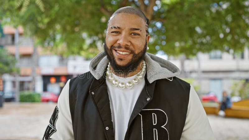 Un sonriente negro barbudo con perlas posa casualmente al aire libre en un parque urbano con vegetación al fondo imagenes de archivo