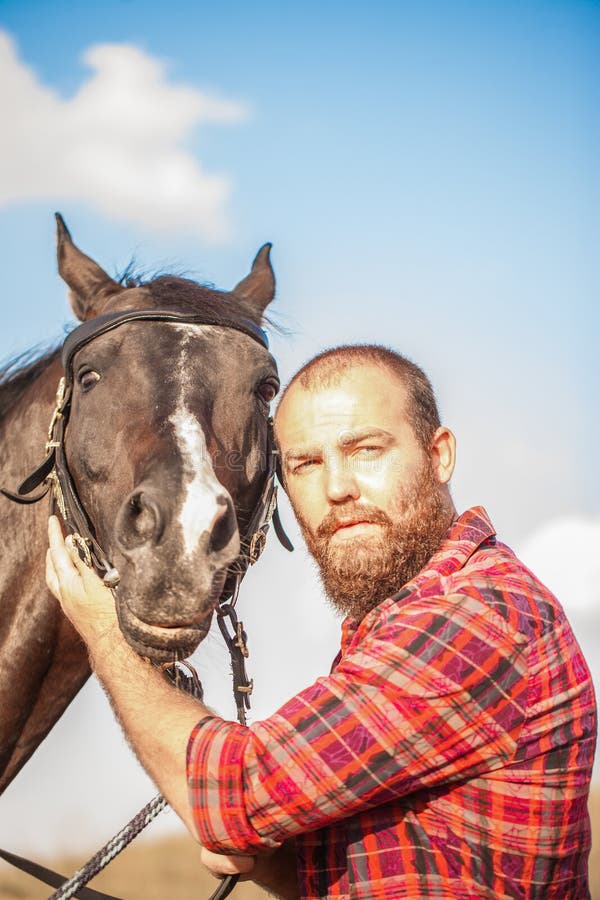 Un tizio brutale con la barba guida un cavallo. agricoltore in camicia di aiuto e. contadino in camicia e stallone nero.