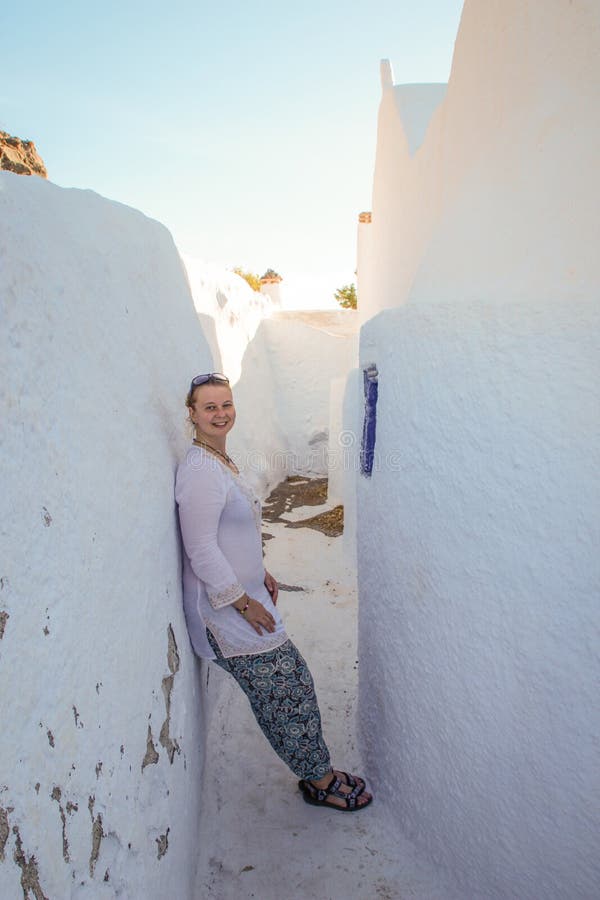 Un turista della ragazza nei sorrisi bianchi dei vestiti e materiali di riempimento sulle pareti bianche della città di Fira immagine stock Un turista della ragazza nei sorrisi bianchi dei vestiti e materiali di riempimento sulle pareti bianche della città di Fira immagine stock
