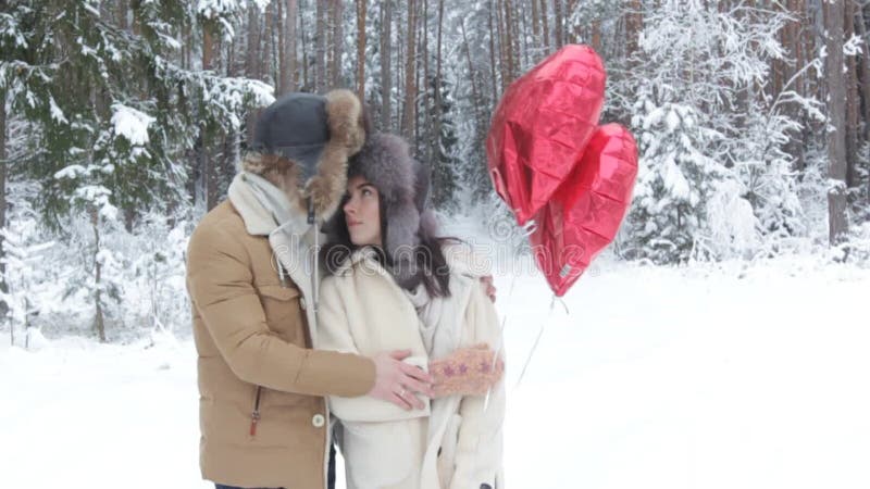Un type doucement embrasse une fille dans une forêt d'hiver neigeuse tenant les boules rouges en forme de coeur. clips vidéos