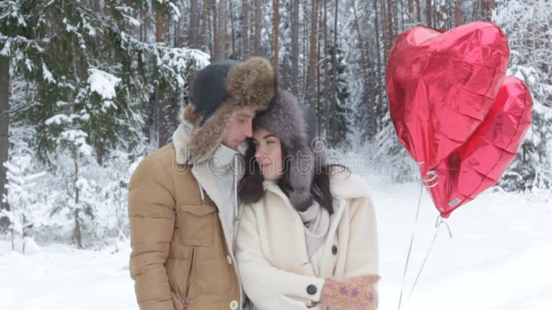Un type embrasse doucement une fille dans une forêt d'hiver enneigée tenant des boules rouges en forme de coeur. clips vidéos