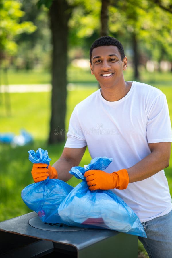 Un type souriant devant la caméra tenant des sacs à déchets. Jour chanceux. un gars à la peau foncée avec des gants de protection souriant à la caméra tenant des sacs de déchets au-dessus de poubelle dans le parc vert