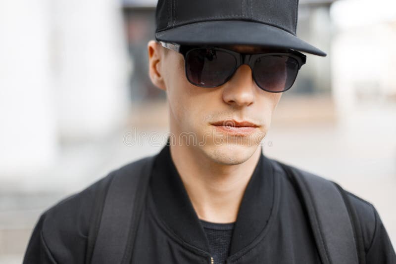 Un tânăr stilat, un hipster într-un baschetbalist negru într-o jachetă elegantă de toamnă neagră în ochelari de soare stă în picio stock images
