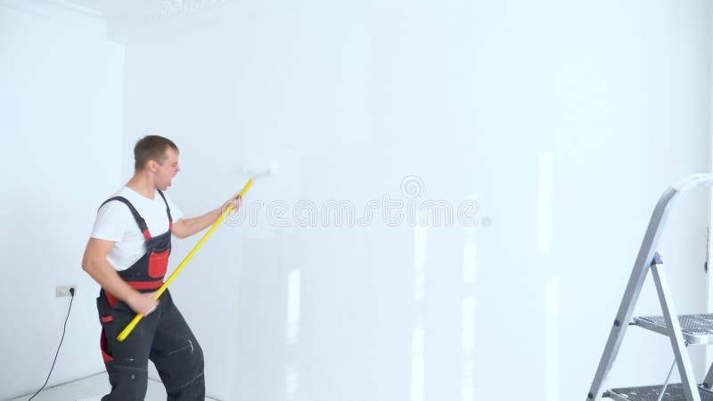 Un uomo allegro in tuta da lavoro dipinge il muro con un rullo mentre danza