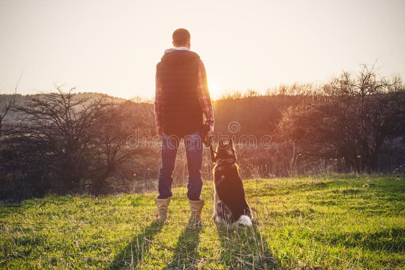 Un uomo con una barba che cammina il suo cane nella natura, stante con una lampadina al sol levante, fondente un'incandescenza ca immagini stock libere da diritti Un uomo con una barba che cammina il suo cane nella natura, stante con una lampadina al sol levante, fondente un'incandescenza ca immagini stock libere da diritti