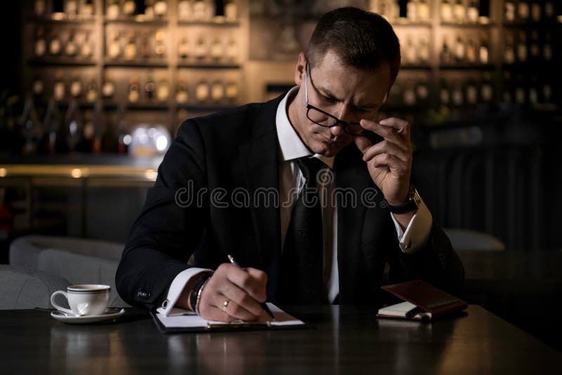 Un uomo d'affari elegante, serio e concentrato che si siede nel ristorante moderno e che fa le note in suo taccuino immagini stock libere da diritti Un uomo d'affari elegante, serio e concentrato che si siede nel ristorante moderno e che fa le note in suo taccuino immagini stock libere da diritti