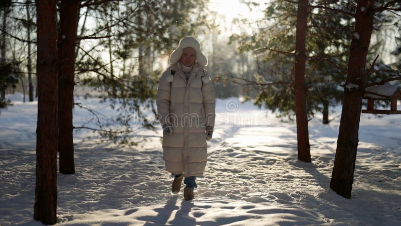 Un uomo felice con una giacca bianca sta camminando in un parco di neve d'inverno tra alberi nudi stock footage