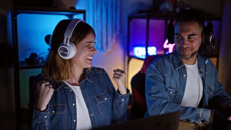 Un uomo gioioso e una donna che indossano cuffie in una stanza da gioco con luci al neon competono in un videogioco di notte