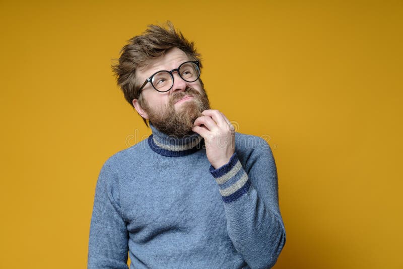 Shaggy pensive man in glasses and an old sweater is trying to remember something by touching his beard with hand. Isolated on yellow background. Shaggy pensive man in glasses and an old sweater is trying to remember something by touching his beard with hand. Isolated on yellow background..