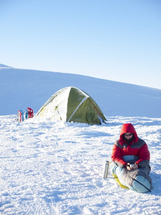 Un uomo si siede in un sacco a pelo vicino alla tenda ed alle racchette da neve immagine stock Un uomo si siede in un sacco a pelo vicino alla tenda ed alle racchette da neve immagine stock