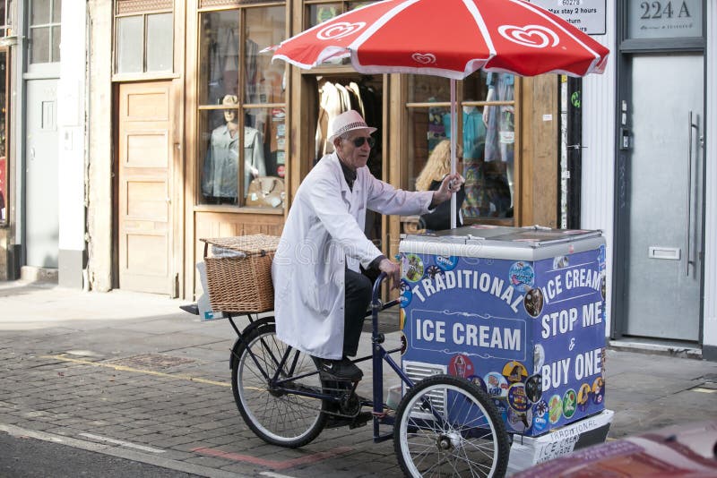 Un vendeur plus âgé de crème glacée dans une robe longue blanche monte une bicyclette avec son chariot de crème glacée image libre de droits Un vendeur plus âgé de crème glacée dans une robe longue blanche monte une bicyclette avec son chariot de crème glacée image libre de droits