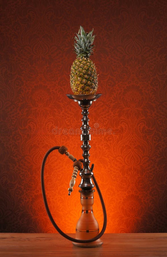 A beautiful old blue ceramic hookah with a fresh pineapple on top standing on a wooden desk. The image is taken in a studio on an orange vintage background. A beautiful old blue ceramic hookah with a fresh pineapple on top standing on a wooden desk. The image is taken in a studio on an orange vintage background.