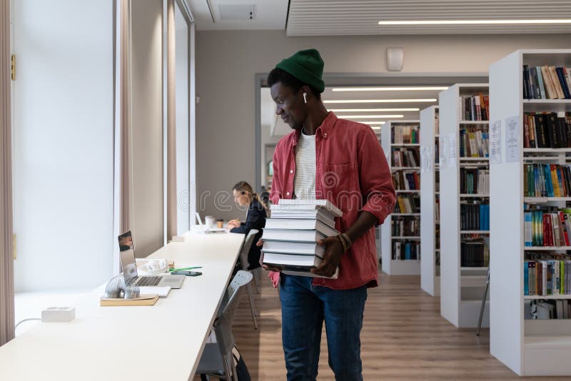 Un étudiant africain tenant une pile de livres et regardant l'écran d'ordinateur portable en étudiant à la bibliothèque. Bibliothèques et apprentissage en ligne. un étudiant africain américain tient une pile de livres et regarde l'écran d'ordinateur portable en étudiant à la bibliothèque universitaire. environnement d'apprentissage et concept d'éducation