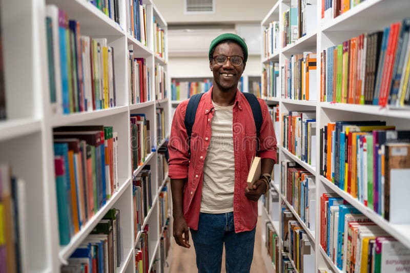 Un étudiant américain souriant tient un livre dans une bibliothèque universitaire entre bibliothèques. photos libres de droits