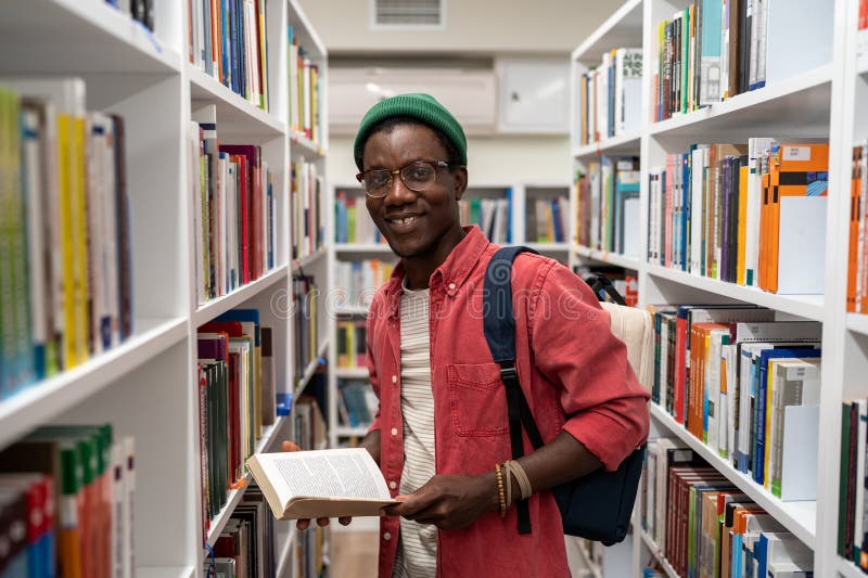 Un étudiant américain souriant tient un livre dans une bibliothèque universitaire entre bibliothèques. photographie stock libre de droits