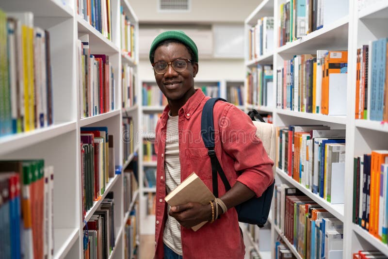 Un étudiant américain souriant tient un livre dans une bibliothèque universitaire entre bibliothèques. photo stock