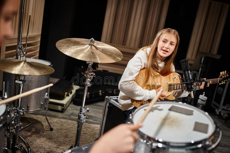 adorable blonde teenage girl in casual attire playing guitar and looking at her friend playing drums, stock photo. adorable blonde teenage girl in casual attire playing guitar and looking at her friend playing drums, stock photo