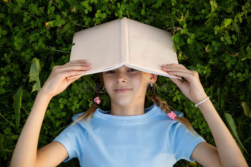 One teen girl student lying with book on green grass lawn in park, studies, writes, reads. One teen girl student lying with book on green grass lawn in park, studies, writes, reads.