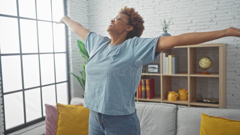 Una alegre afroamericana que se extiende en una sala de estar moderna con grandes ventanas que evocan el frescor y la relajación imagen de archivo libre de regalías
