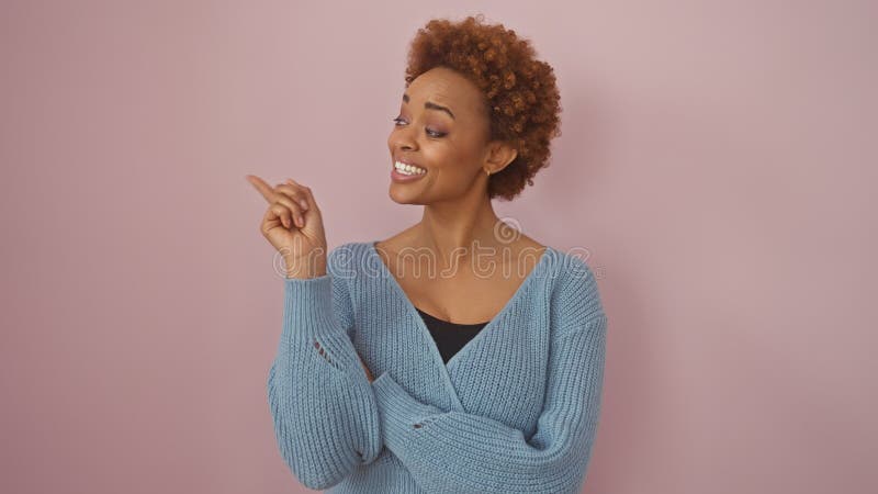 Una alegre mujer afroamericana en un suéter señala alegremente a un fondo rosado que encarna positividad y confianza foto de archivo