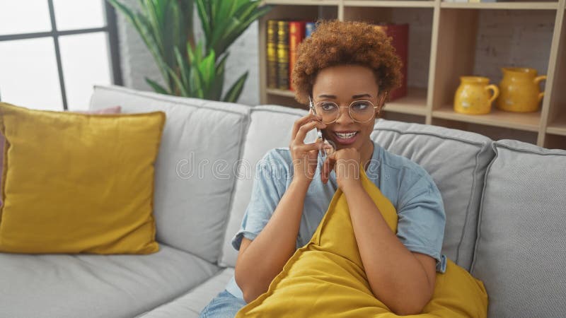 Una atractiva afroamericana que disfruta de una conversación por teléfono cómodamente en su elegante sala de estar foto de archivo