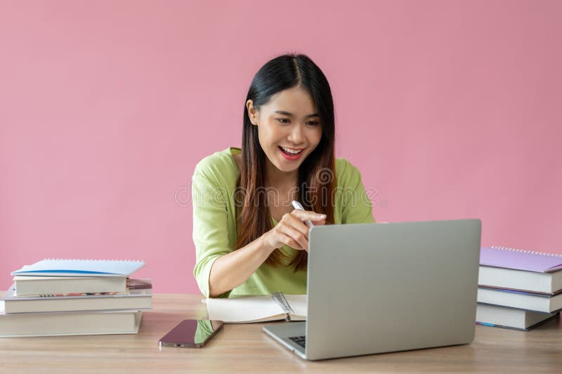 Una atractiva estudiante universitaria asiática está disfrutando de una clase en línea en su laptop. Una atractiva y alegre joven estudiante universitaria asiática está disfrutando de una clase en línea en su laptop en la mesa de estudio. fondo rosa aislado. elearning online study