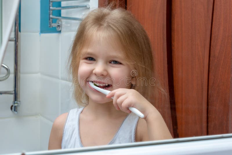 Una bambina con capelli biondi che puliscono i suoi denti Il bambino sta sorridendo alla riflessione nello specchio immagini stock Una bambina con capelli biondi che puliscono i suoi denti Il bambino sta sorridendo alla riflessione nello specchio immagini stock