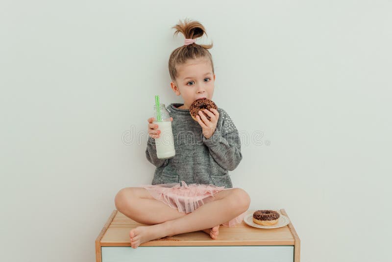 Una bambina sorridente è seduta sulla commodalità con una ciambella di cioccolato e una bottiglia di latte. ritratto di una ragazz fotografia stock