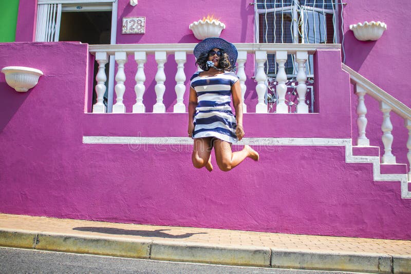 Una bella donna africana in un vestito a strisce blu e bianco che salta davanti ad una casa tradizionale con le pareti rosa La BO immagine stock libera da diritti Una bella donna africana in un vestito a strisce blu e bianco che salta davanti ad una casa tradizionale con le pareti rosa La BO immagine stock libera da diritti
