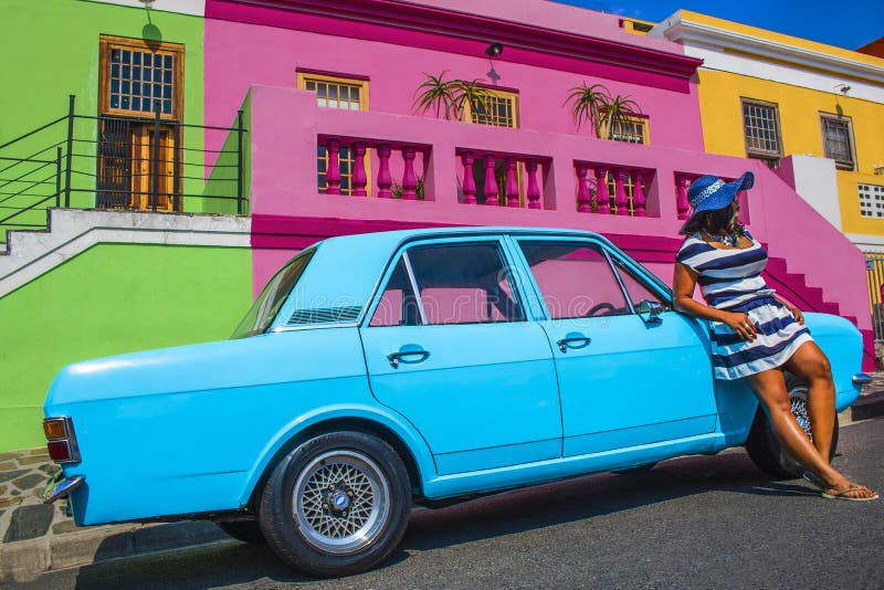 Una bella donna africana in un vestito a strisce blu e bianco davanti a Ford Cortina d'annata ed alle case tradizionali della BO- fotografia stock Una bella donna africana in un vestito a strisce blu e bianco davanti a Ford Cortina d'annata ed alle case tradizionali della BO- fotografia stock