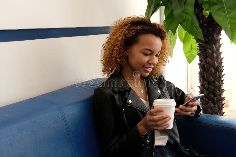 Una bella giovane donna di colore moderna, in un bomber con i airpods in suo orecchio, si siede su un sofà blu La ragazza accanto fotografie stock libere da diritti Una bella giovane donna di colore moderna, in un bomber con i airpods in suo orecchio, si siede su un sofà blu La ragazza accanto fotografie stock libere da diritti