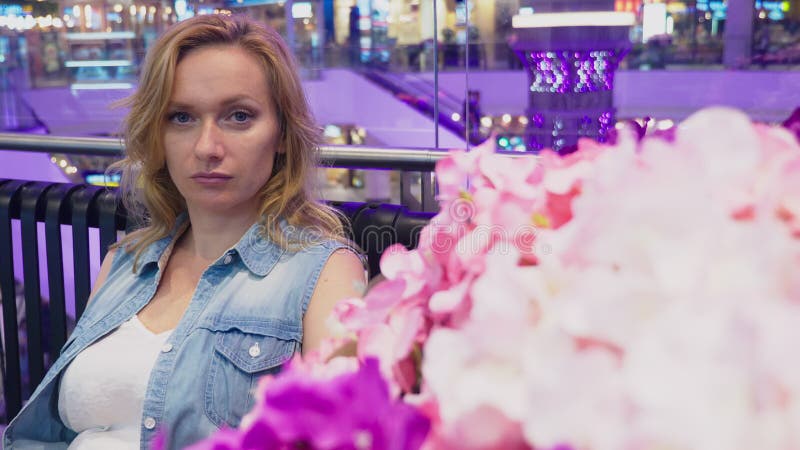 Una bella giovane donna sta sedendosi su un banco in un grande centro commerciale moderno vicino ad un letto di fiore fotografia stock Una bella giovane donna sta sedendosi su un banco in un grande centro commerciale moderno vicino ad un letto di fiore fotografia stock