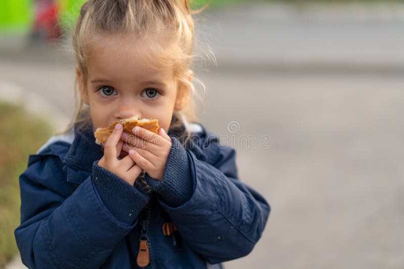 Una bellissima ragazzina caucasica dai capelli biondi e che mangia pane avidamente con le mani guarda la telecamera con occhi tri
