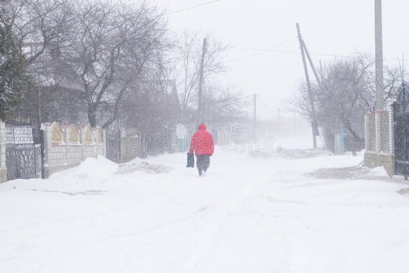 Una calle nevosa del pueblo durante una tormenta de la nieve y un hombre en una chaqueta roja fotos de archivo libres de regalías Una calle nevosa del pueblo durante una tormenta de la nieve y un hombre en una chaqueta roja fotos de archivo libres de regalías