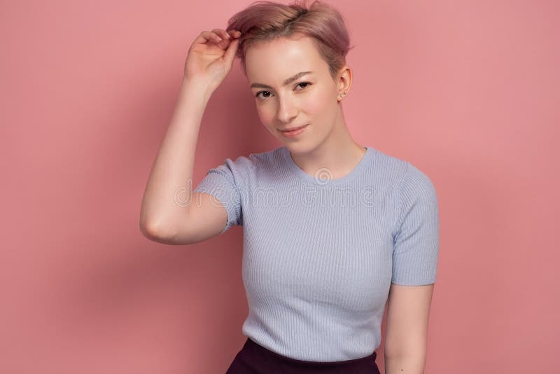 Una chica con el pelo rosado corto en una camiseta se pone de pie sobre un fondo rosado, mirando la cámara y alisando su pelo fotografía de archivo libre de regalías Una chica con el pelo rosado corto en una camiseta se pone de pie sobre un fondo rosado, mirando la cámara y alisando su pelo fotografía de archivo libre de regalías