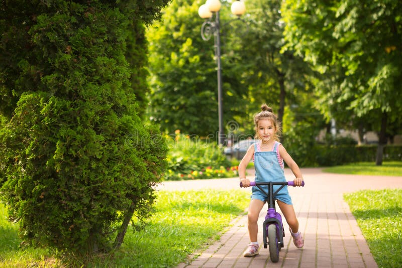 Una Chica Con Traje De Salto De Denim Monta En Una Bicicleta Corriendo En Verano En Un Parque Verde. Niños Scooter Activo Foto de archivo - Imagen equilibrio, paseos: 209462018