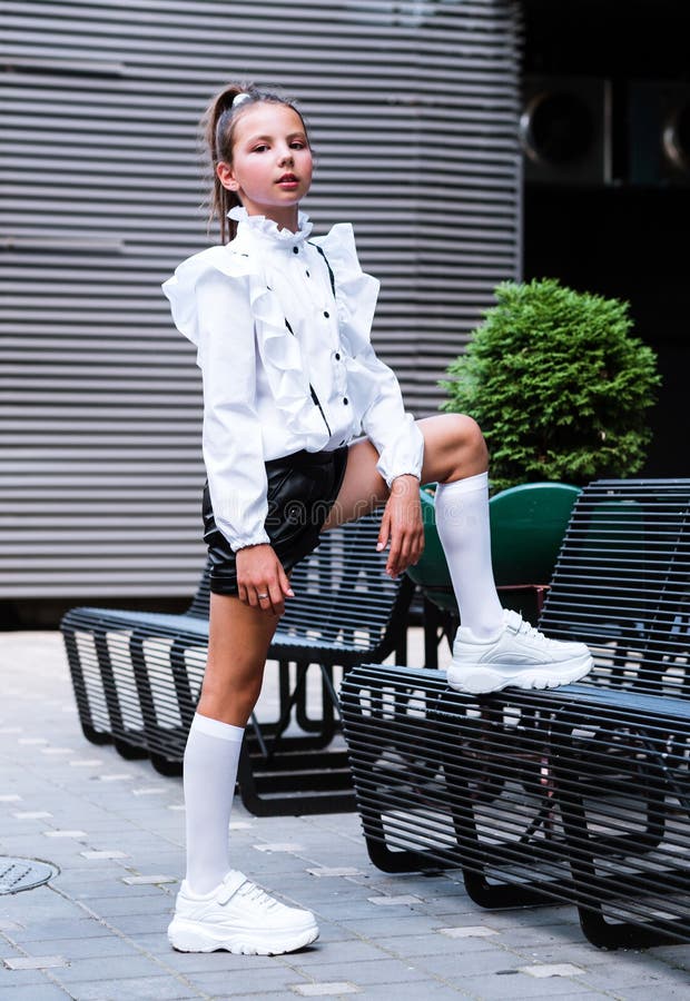 A fashionable girl in a school uniform, socks and athletic shoes posing with her foot on an iron bench. Vertical photo. A fashionable girl in a school uniform, socks and athletic shoes posing with her foot on an iron bench. Vertical photo