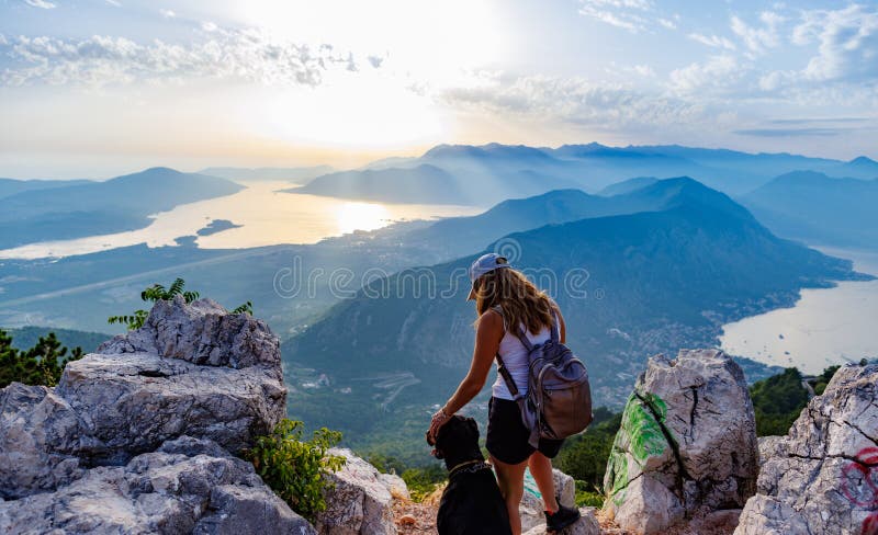 A young happy girl with a backpack stands with her faithful dog Rottweiler on top of a high Montenegrin mountain against the backdrop of coastal cities, the Adriatic Sea and a bright sunny sky. A young happy girl with a backpack stands with her faithful dog Rottweiler on top of a high Montenegrin mountain against the backdrop of coastal cities, the Adriatic Sea and a bright sunny sky