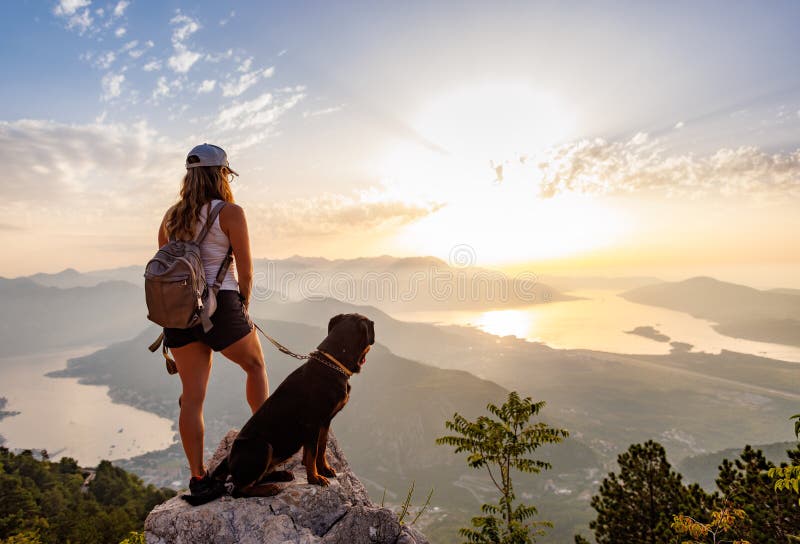 A young happy girl with a backpack stands with her faithful dog Rottweiler on top of a high Montenegrin mountain against the backdrop of coastal cities, the Adriatic Sea and a bright sunny sky. A young happy girl with a backpack stands with her faithful dog Rottweiler on top of a high Montenegrin mountain against the backdrop of coastal cities, the Adriatic Sea and a bright sunny sky