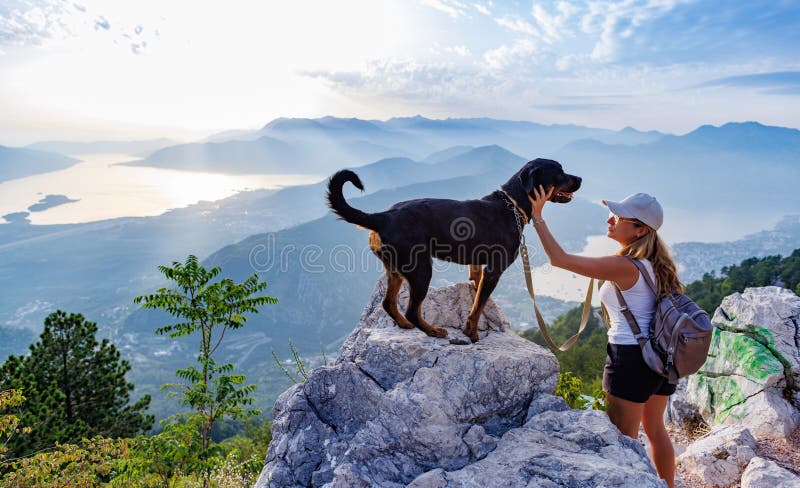 A young happy girl with a backpack stands with her faithful dog Rottweiler on top of a high Montenegrin mountain against the backdrop of coastal cities, the Adriatic Sea and a bright sunny sky. A young happy girl with a backpack stands with her faithful dog Rottweiler on top of a high Montenegrin mountain against the backdrop of coastal cities, the Adriatic Sea and a bright sunny sky