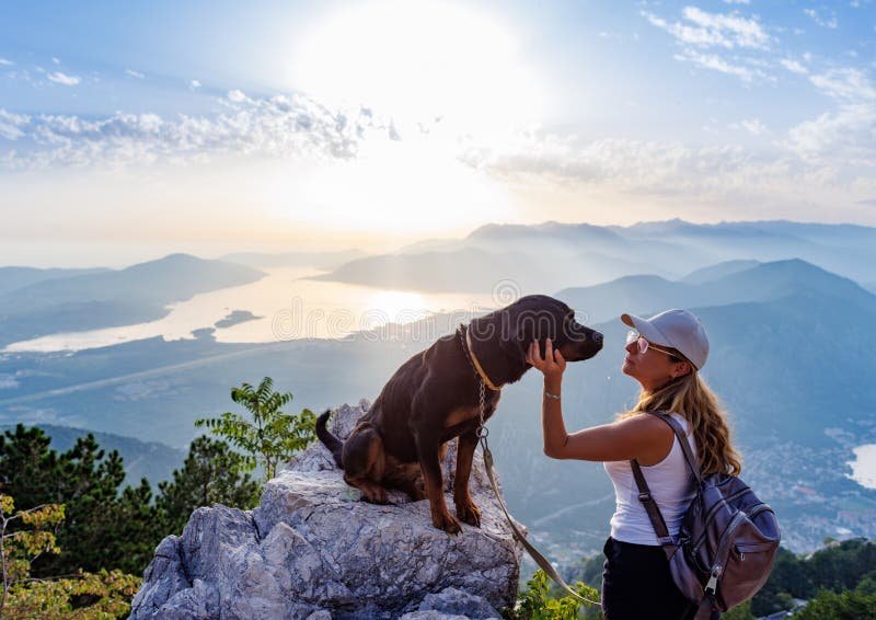 A young happy girl with a backpack stands with her faithful dog Rottweiler on top of a high Montenegrin mountain against the backdrop of coastal cities, the Adriatic Sea and a bright sunny sky. A young happy girl with a backpack stands with her faithful dog Rottweiler on top of a high Montenegrin mountain against the backdrop of coastal cities, the Adriatic Sea and a bright sunny sky