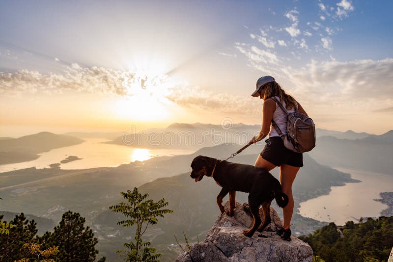 A young happy girl with a backpack stands with her faithful dog Rottweiler on top of a high Montenegrin mountain against the backdrop of coastal cities, the Adriatic Sea and a bright sunny sky. A young happy girl with a backpack stands with her faithful dog Rottweiler on top of a high Montenegrin mountain against the backdrop of coastal cities, the Adriatic Sea and a bright sunny sky